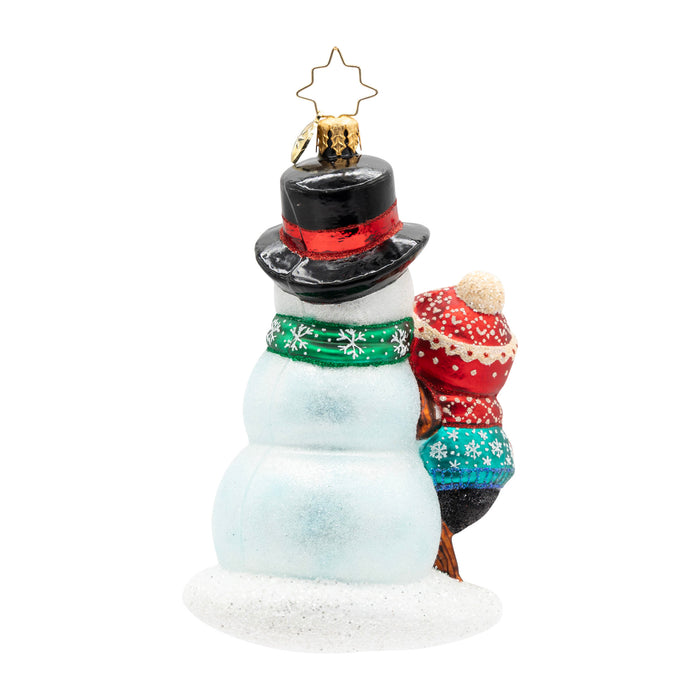 Back view of a penguin ornament wearing a blue coat and a matching red hat and scarf, finishing decorating the snowman it built.