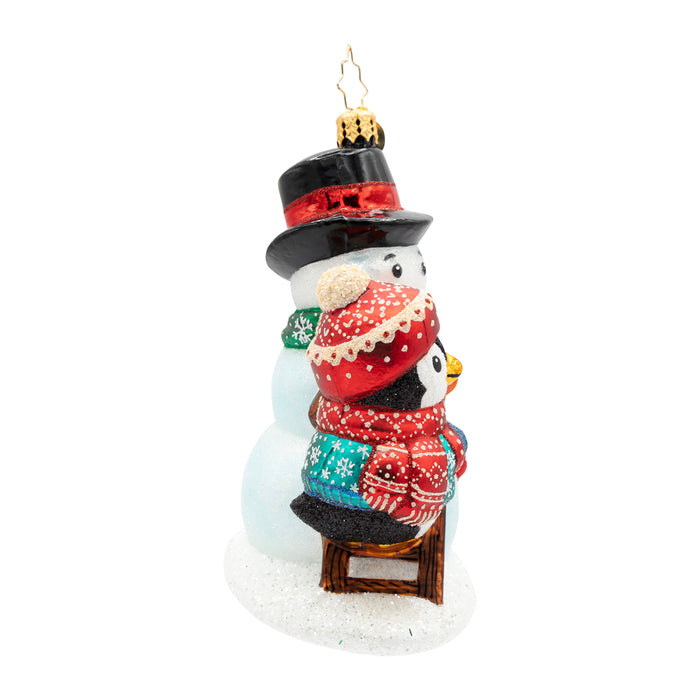 Side view of a penguin ornament wearing a blue coat and a matching red hat and scarf, finishing decorating the snowman it built. Showcases the little stool the penguin stands on.