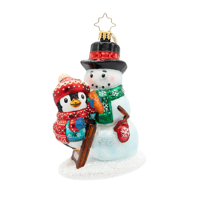 Front view of a penguin ornament wearing a blue coat and a matching red hat and scarf, finishing decorating the snowman it built.