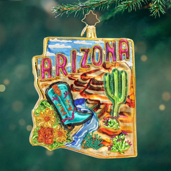 Ornament shaped in the state of Arizona featuring a blue sky with the Grand Canyon, turquois boots, catus, and the word Arizona in pink and gold trim. View of front
