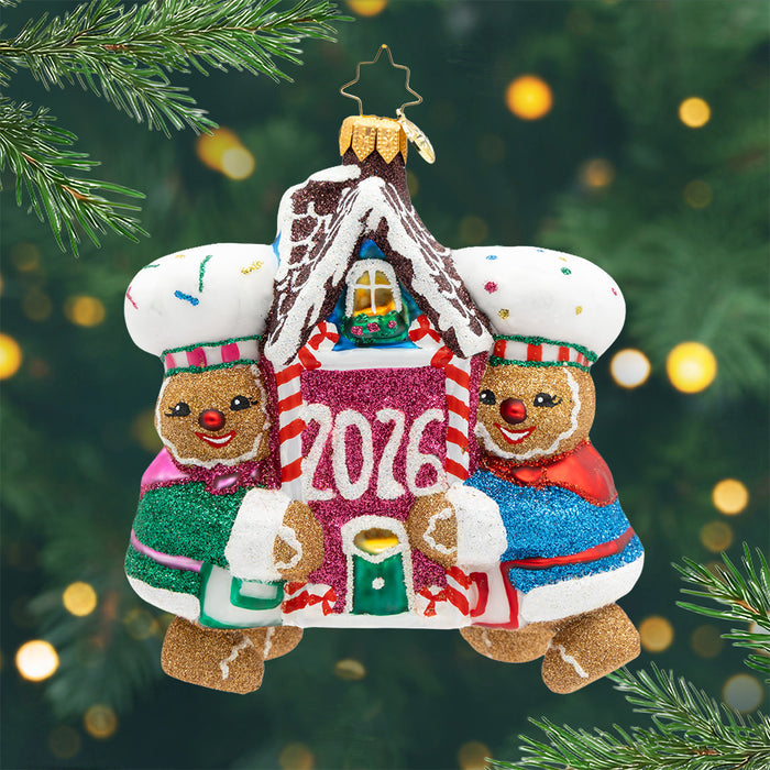 Baking Up Joy 2026. A sparkling Ornament of two gingerbread in blue and green coats and holding a gingerbread house with pink in the front and the year 2026. View of front