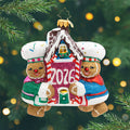 Baking Up Joy 2026. A sparkling Ornament of two gingerbread in blue and green coats and holding a gingerbread house with pink in the front and the year 2026. View of front