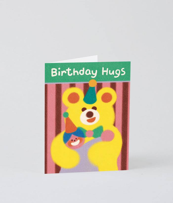 a big birthday bear hug on this stripy birthday card for kids. a big yellow bear hugs a person in a party hat and it says Birthday Hugs on a green background.