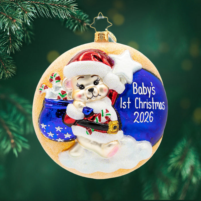 Darling 1st Christmas Personalized. Round ornament of a bear in a red Santa suit, holding a starry blue gift bad. on top of a glittery moon. View of front