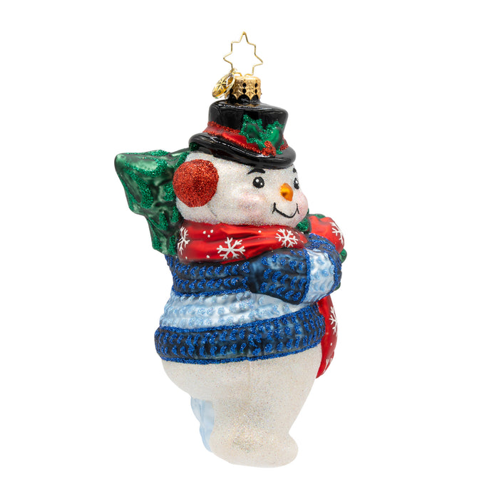 Side view of a decorative Christmas ornament shaped like a snowman holding a small tree, wearing a blue sweater and red mittens.