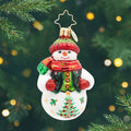 Festive Folk Flair Gem. Ornament of a snowman in a black and green vest with a red and green sparkly hat and matching scarf, along with a tree painted on the bottom. View of front