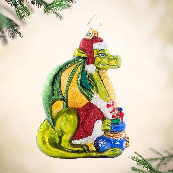 Fire-Breathing Friend. Ornament of a giant green dragon wearing a sparkling Santa suit and holding a giant blue gift bag. View of front