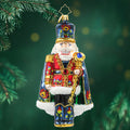 Nutcracker ornament dressed in a red suit with matching blue hat, cape, and boots, each with floral patterns as well as gold detailing and trim. View of front