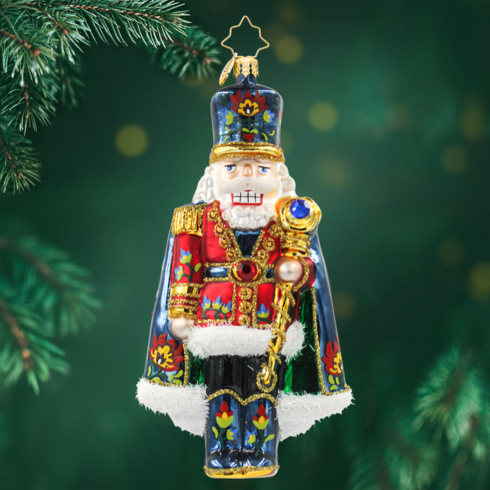Nutcracker ornament dressed in a red suit with matching blue hat, cape, and boots, each with floral patterns as well as gold detailing and trim. View of front