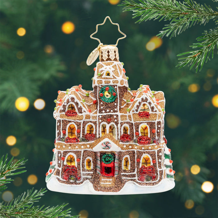 Gingerbread Manor Gem. Ornament of a gingerbread house in a sparkling brown with golden windows and red bows in front of them and a red door. View of front.
