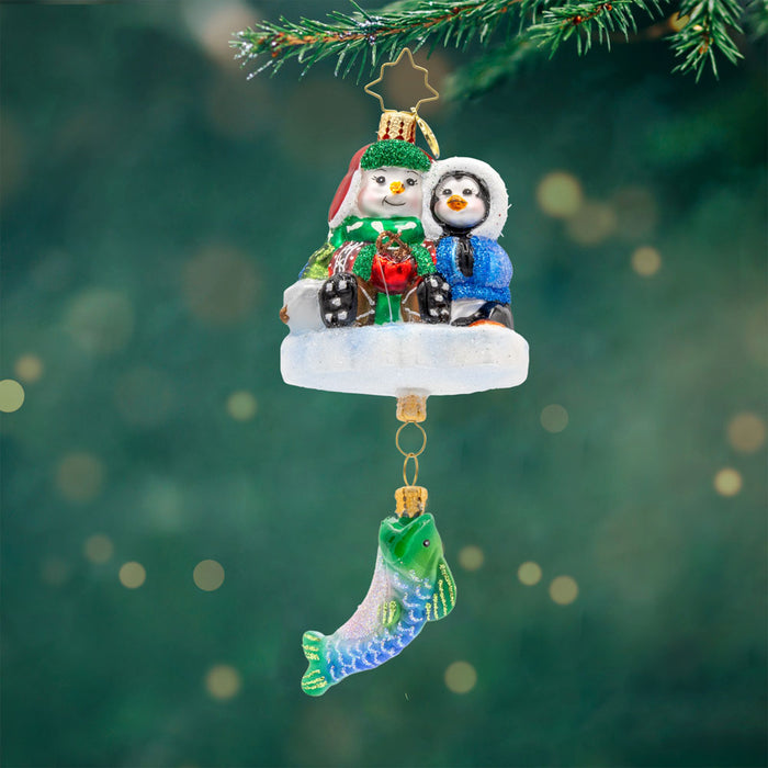 Ice Fishing Snow Pals Gem ornament. A snowman and penguin sit bundled up on a block of ice with a fishing pole. A fish dangles below. View of front.