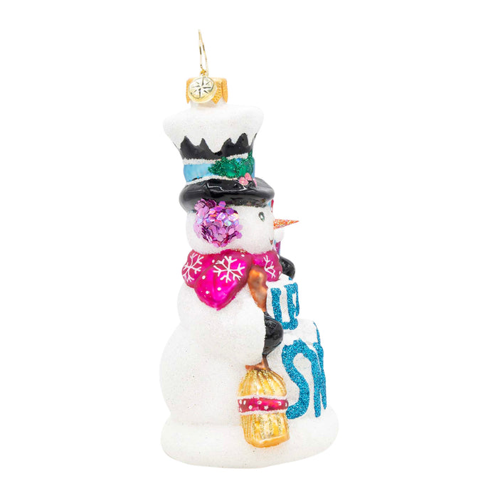 Side of a decorative snowman ornament holding a broom, with the phrase "Let It Snow" in blue on the front. Purple earmuffs and pink scarf with white snowflake designs.