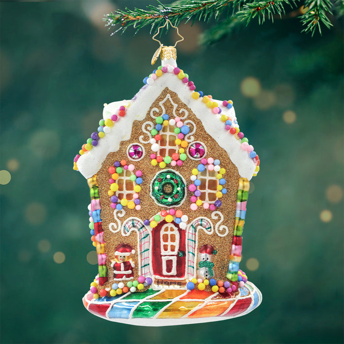 House shaped ornament adorned with details like gumdrops, candy canes, and frosting with colors of brown, white, pink, blue, green and yellow. View of front