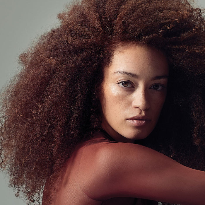 Model with voluminous, defined brown curls wearing a rust-coloured top against a muted grey-green studio background.