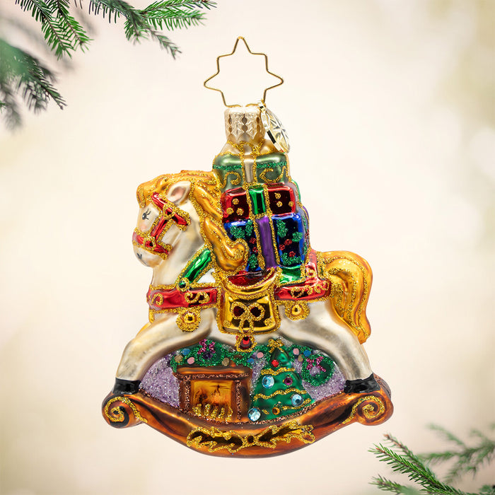 Rockin' Around Christmas Gem. Sparkling ornament of a white rocking horse with a golden mane holding colorful presents of red, blue and green with a brown fireplace and a decorated Christmas tree beneath it. View of front
