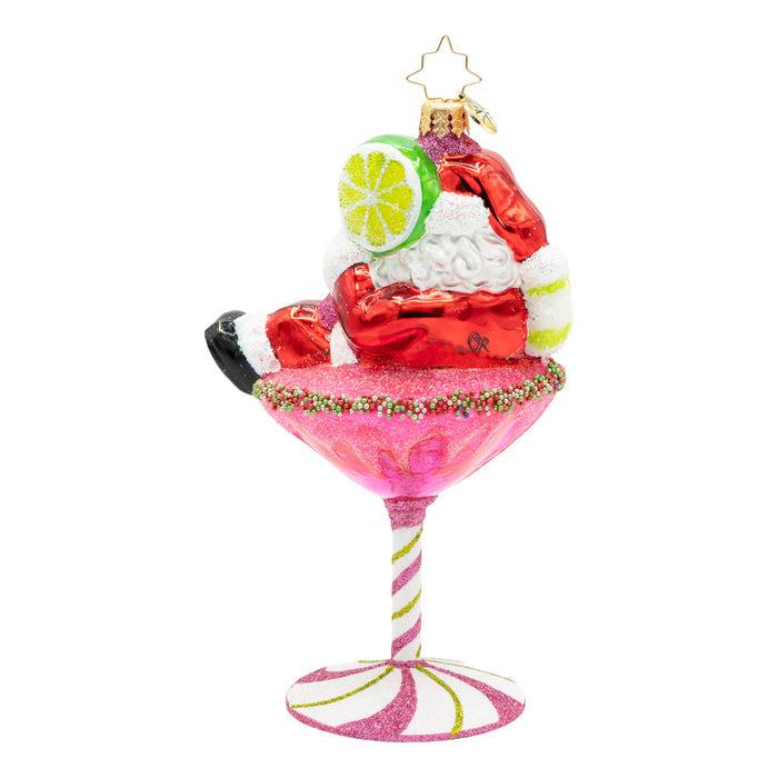 Back view of ornament of Santa laying on top of a pink martini drink with a base of a pink peppermint, while holding a lime.