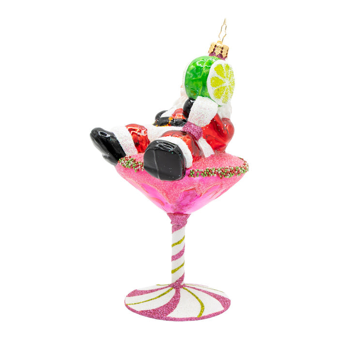 Side view of ornament of Santa laying on top of a pink martini drink with a base of a pink peppermint, while holding a lime.