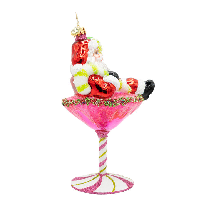 Side view of ornament of Santa laying on top of a pink martini drink with a base of a pink peppermint, while holding a lime.