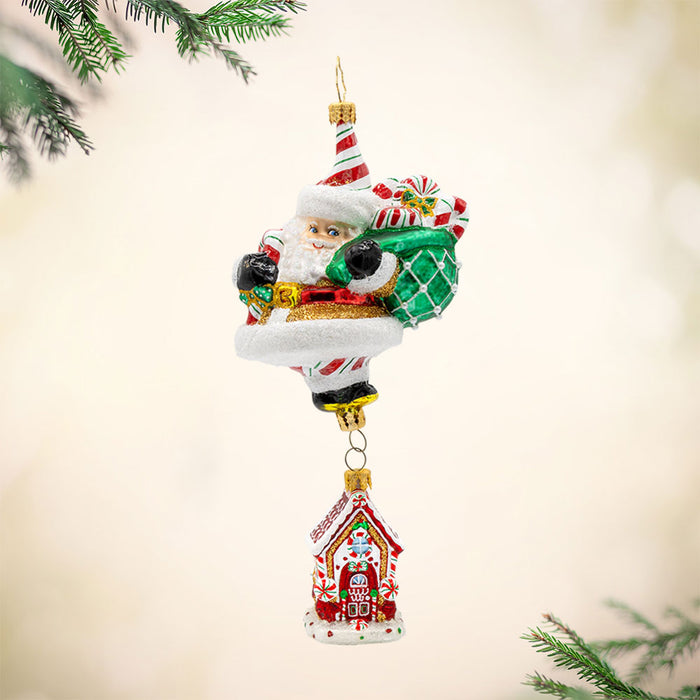 Ornament features a gingerbread house dangling from Santa's feet. Santa is holding a bag overflowing with treats. View of front.