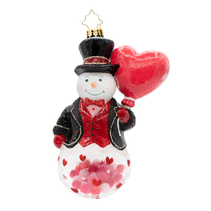 Front view of a snowman ornament wearing a red and black tuxedo holding a red heart. The base of the ornament has red and pink confetti on the inside. Placed infront of a white background