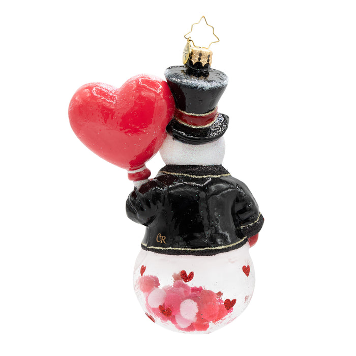 Back view of a snowman ornament wearing a red and black tuxedo holding a red heart. The base of the ornament has red and pink confetti on the inside. Placed infront of a white background