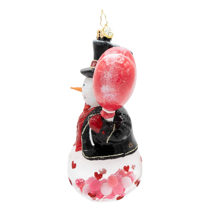 Side view of a snowman ornament with a top hat, black coat, and red heart design on the base of the ornament. showcases the heart shaped balloon. Placed on a white background