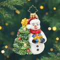 Snowy & Bright! Gem. Snowman ornament dressed in a red hat and scarf, holding a blue present next to a decorated Christmas tree. View of front