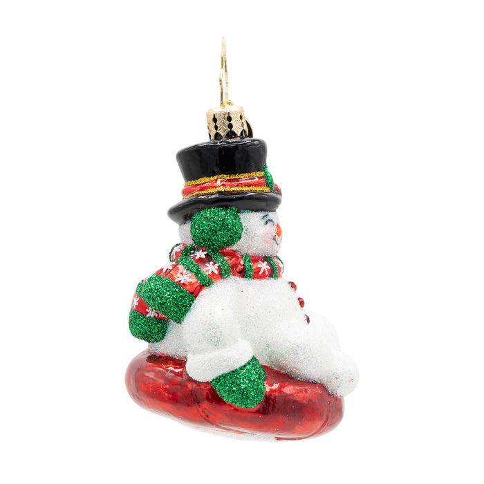 Side view of ornament of a snowman wearing a red and green scarf on a red sled.