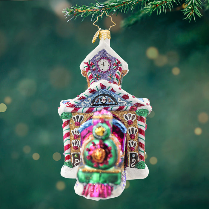 An ornament in the shape of a train station with shimmering colors in purple, blue green, and pink. View of front